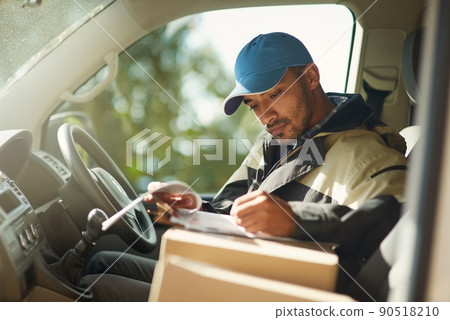 Updating his delivery status. Shot of a delivery man reading addresses while sitting in a delivery van. Updating his delivery status. Shot of a delivery man reading addresses while sitting in a delivery van. 90518210