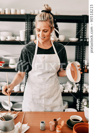 I love painting my pottery. Cropped shot of an attractive mature woman standing alone and painting a pottery bowl in her workshop. 90518215