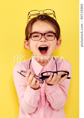 Shes excited about spectacles. A cute little girl standing against a yellow background with a hand full of glasses. 90519960