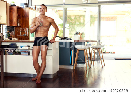 I love my time off just to enjoy a coffee. Shot of a handsome young shirtless man drinking a cup of coffee in the kitchen at home. 90520229