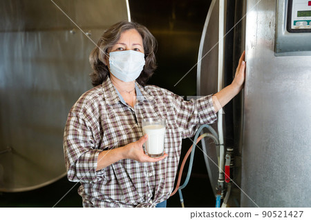 Mature woman in protective mask is holding glass of milk at cow farm Mature woman in protective mask is holding glass of milk at cow farm 90521427