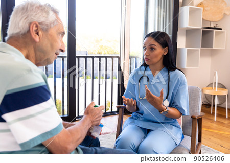 Biracial female physiotherapist talking with caucasian senior man while sitting on chair at home 90526406