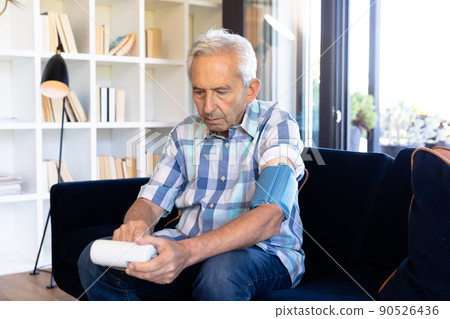 Senior caucasian man examining blood pressure with blood pressure monitor while sitting on sofa 90526436