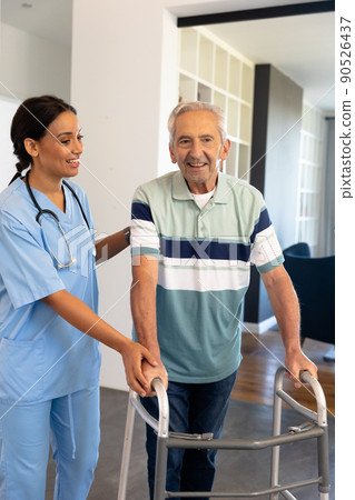 Smiling multiracial female physiotherapist assisting senior man in walking with walker at home 90526437