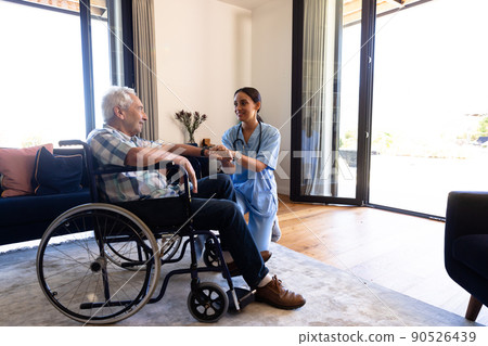 Biracial female physiotherapist talking and holding senior man's hand sitting on wheelchair at home 90526439