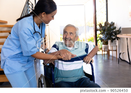 Biracial female physiotherapist assisting caucasian senior man in getting up from wheelchair at home 90526450