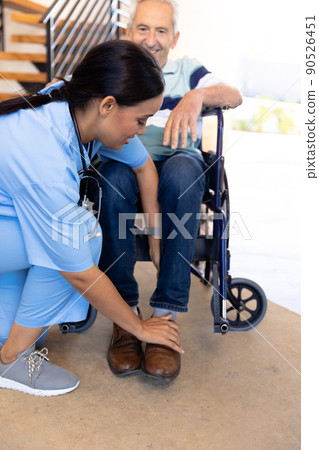 Biracial female physiotherapist adjusting legs of caucasian senior man sitting on wheelchair at home 90526451
