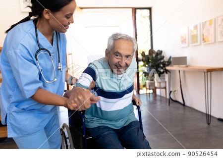 Biracial female physiotherapist assisting smiling caucasian senior man in getting up from wheelchair 90526454