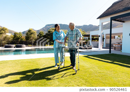 Biracial female doctor assisting caucasian senior man in walking with walker in yard on sunny day 90526461