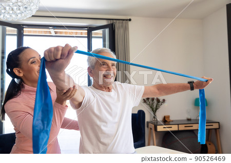 Biracial female physiotherapist assisting caucasian senior man stretching resistance band at home 90526469