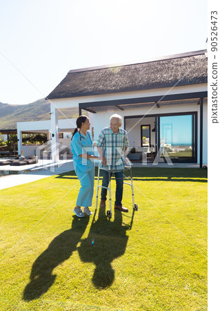 Smiling biracial female doctor assisting caucasian senior man in walking with walker in yard Smiling biracial female doctor assisting caucasian senior man in walking with walker in yard 90526473