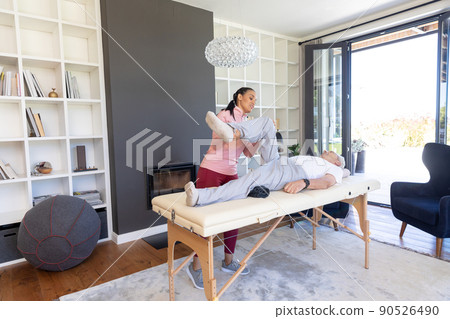 Biracial female physiotherapist stretching leg of caucasian senior man lying on table at home 90526490