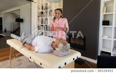 Biracial female physiotherapist gesturing while guiding caucasian senior man lying on table at home 90526491