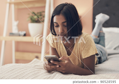 What would quarantine be without your social connection. Shot of a young woman using a smartphone while relaxing on her bed at home. What would quarantine be without your social connection. Shot of a young woman using a smartphone while relaxing on her bed at home. 90528595