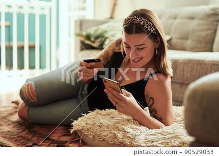 Shopping just gets easier and easier. Shot of a young woman using a smartphone and credit card in her living room at home. Shopping just gets easier and easier. Shot of a young woman using a smartphone and credit card in her living room at home. 90529033