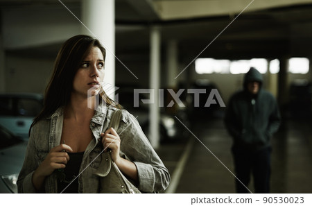I think that guy is following me.... A young woman in a parking lot looking concerned as someone follows her. 90530023