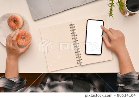 Top shot of a female student using smartphone and eating doughnut in her workspace. 90534104