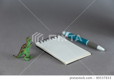 Dinosaur miniature, notepad and ballpoint pen against a gray background. Small green figure of an animal of prey standing on its hind legs. Spring-loaded caged notebook Dinosaur miniature, notepad and ballpoint pen against a gray background. Small green figure of an animal of prey standing on its hind legs. Spring-loaded caged notebook 90537833
