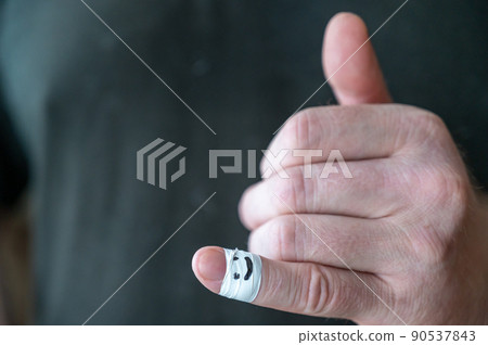 The man shows his hand a thumbs-up gesture. The first phalanx of his little finger is wrapped in white ribbon. A smiling face is painted on the bandage. Positive emotion concept. Selective Focus. 90537843