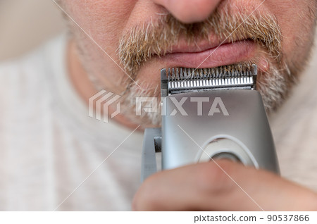 A mature man shaving. The bottom of a man's face. Man cuts facial hair with a hair clipper. A male with stubble. Gray hair on his beard and mustache. Close-up. Selective focus. 90537866