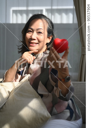 Middle aged woman holding heart shape and smiling at camera. Elderly care, insurance, heart health and quality of life for elderly concept Middle aged woman holding heart shape and smiling at camera. Elderly care, insurance, heart health and quality of life for elderly concept 90537904
