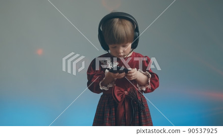 Cute little girl in big headphones and a beautiful vintage dress with lace is listening to music on her phone. There is a color music in the background. The concept of music mania and audiophilia 90537925