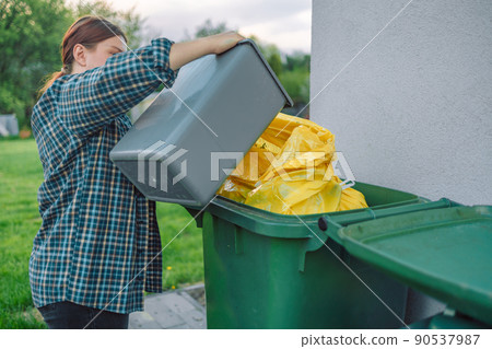 European 30s woman throwing garbage into the recycling bin in the backyard near the house. Separating recyclable garbage plastic European 30s woman throwing garbage into the recycling bin in the backyard near the house. Separating recyclable garbage plastic 90537987