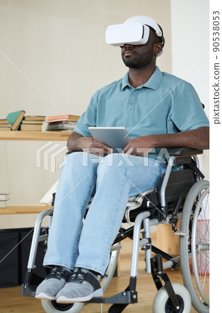 African AMerican man in VR goggles sitting in wheelchair using digital tablet for virtual reality therapy 90538053