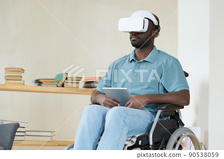 African young man sitting in wheelchair with digital tablet and connecting VR goggles for playing game online 90538059