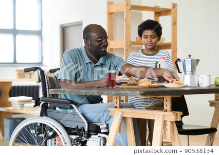 African single father sitting in wheelchair at table and having breakfast with his son in the morning at home 90538134