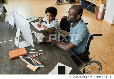 African disabled father sitting in wheelchair using computer at table to help his son with homework African disabled father sitting in wheelchair using computer at table to help his son with homework 90538191
