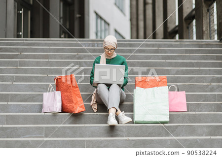 Muslim lady in hijab sitting on the steps of a large shopping mall and ordering clothes online Muslim lady in hijab sitting on the steps of a large shopping mall and ordering clothes online 90538224