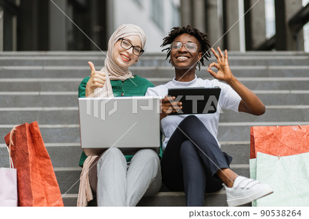 African and Muslim women sitting on the steps of mall with laptop and tablet and shopping online. African and Muslim women sitting on the steps of mall with laptop and tablet and shopping online. 90538264