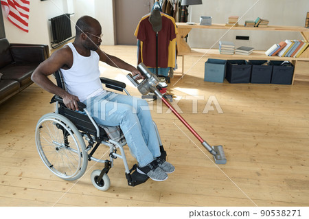 African man with disability sitting in wheelchair and cleaning floor in room using vacuum cleaner without help 90538271