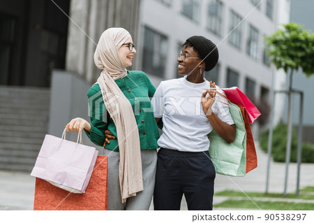 Two multiethnical girls, walking down the old city street after shopping, carrying colorful bags Two multiethnical girls, walking down the old city street after shopping, carrying colorful bags 90538297