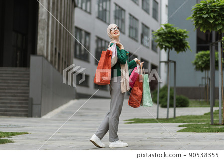 Muslim woman, wearing grey pants and green jacket, walking at the street with colorful shopping bags 90538355