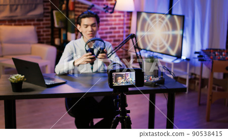 Asian man reviewing headphones product on camera, filming video for podcast episode on social media. Live broadcasting vlog to recommend earphones, show production at web station. 90538415