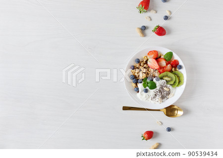 White ceramic bowl of homemade granola with yogurt, fresh berries and fruits on white wooden table background top view. White ceramic bowl of homemade granola with yogurt, fresh berries and fruits on white wooden table background top view. 90539434