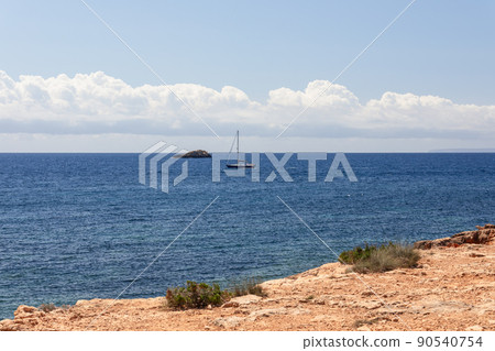 Rocky sandstone coast with sparse vegetation, ripples on sea, sailing yacht with stowed sails. 90540754