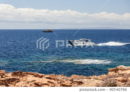 Small silver motor boat moves along the rocky yellow coast of Ibiza island 90540766