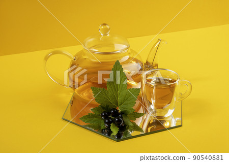 Black currant tea. Transparent glass teapot, cup of tea and black currant berries on a yellow background. Calming drink concept Black currant tea. Transparent glass teapot, cup of tea and black currant berries on a yellow background. Calming drink concept 90540881