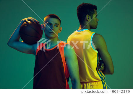 Portrait of two young men, professional basketball players standing back to back, posing isolated over blue studio background in neon light. Team 90542379