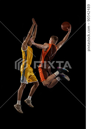 Portrait of two young men, professional basketball players in a jump, throwing ball into basket isolated over black studio background. Winner 90542489