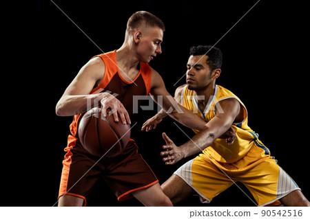 Portrait of two young men, basketball players in uniform training, practising team game isolated over black studio background. Carrying ball. Portrait of two young men, basketball players in uniform training, practising team game isolated over black studio background. Carrying ball. 90542516