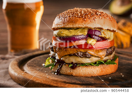 Cheeseburger, made from rye bun with tomato slice and melted cheese on a roasted beef, and lettuce leaf on a wooden table, with a glass of beer 90544458