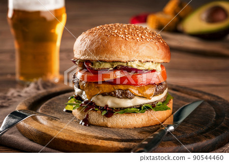 Cheeseburger, made from rye bun with tomato slice and melted cheese on a roasted beef, and lettuce leaf on a wooden table, with a glass of beer Cheeseburger, made from rye bun with tomato slice and melted cheese on a roasted beef, and lettuce leaf on a wooden table, with a glass of beer 90544460