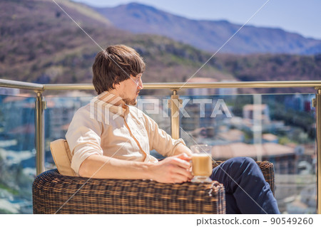 Young man drinking coffee on the balcony, standing behind a mountain Young man drinking coffee on the balcony, standing behind a mountain 90549260