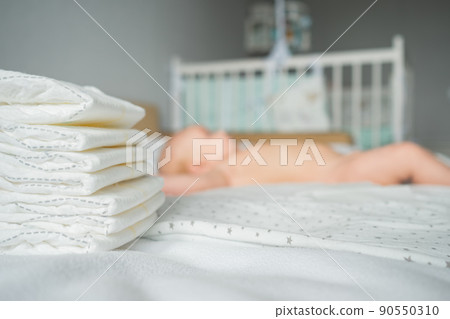 Stack of clean diapers on foreground selective focus and baby girl lying on bed white sheets at home on blurred background. Stack of clean diapers on foreground selective focus and baby girl lying on bed white sheets at home on blurred background. 90550310