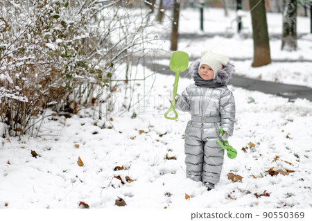 A funny little girl in a warm silver jumpsuit holds a toy shovel. 90550369