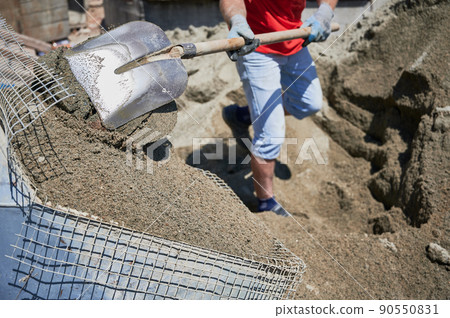 Close up of man with shovel in his hands throwing cementitious floor screed material into container with net. Male in work gloves shoveling sand-cement mix outdoors at construction site. 90550831